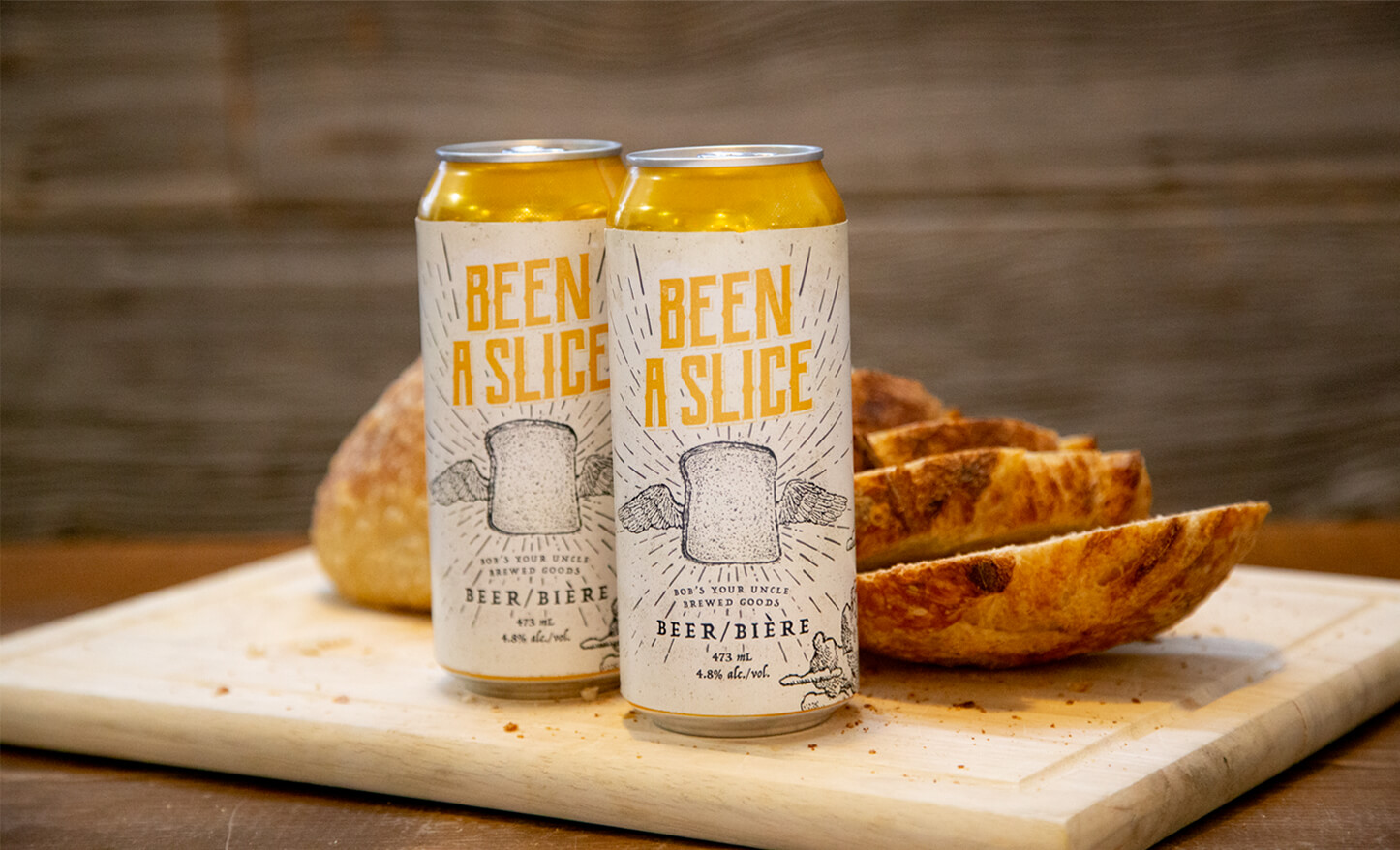 Two cans of Been A Slice beer on a bread board with bread slices in the background.